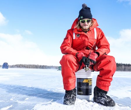 Man in a red snowsuit is sitting on a bucket holding a fishing rod and ice fishing. There is a blue sky behind him.